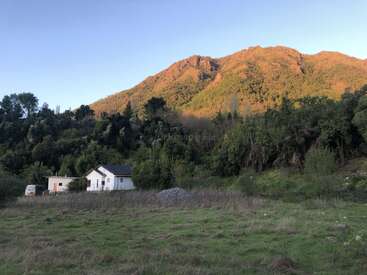 Scène rurale paisible présentant une maison blanche et une camionnette à proximité d'une forêt verte dense, avec une montagne imposante éclairée par le soleil à l'arrière-plan sous un ciel dégagé.