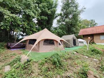 Drei große Zelte sind auf grünen Matten neben einem rustikalen Gebäude und einem Wald aufgebaut. Eine Couch, eine Hängematte, viel Grün und ein wolkenverhangener Himmel sorgen für einen gemütlichen Campingplatz.