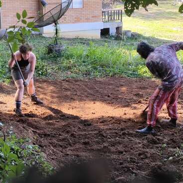 Zwei Menschen arbeiten zusammen in einem Garten und bearbeiten die Erde mit Schaufeln. Im Hintergrund sind ein Backsteinhaus, eine Satellitenschüssel und üppiges Grün zu sehen.