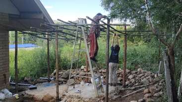 Dos personas construyen una estructura de madera al aire libre. Una está de pie en una escalera trabajando en altura, mientras que la otra ayuda desde el suelo. El entorno es frondoso y verde.