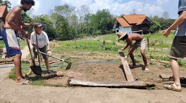 Cuatro personas trabajan juntas al aire libre en un día soleado, preparando la tierra en una zona ajardinada con palas y tablones de madera. Casas rústicas y vegetación les rodean.