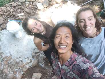 Tres personas sonrientes se toman un selfie al aire libre, de pie sobre un suelo rocoso junto a una estructura de retrete de compost. Parecen felices, posiblemente haciendo voluntariado o trabajando juntos en un proyecto ecológico.