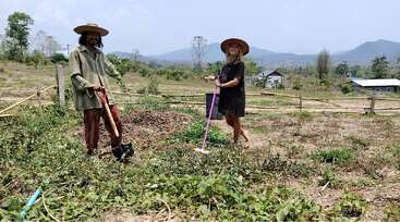 Dos personas están de pie en un campo con palas, sonriendo y llevando sombreros. El fondo muestra montañas, vegetación verde y cielos despejados en un día soleado.