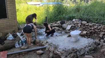 Tres personas trabajan al aire libre, edificando o construyendo una plataforma de piedra y hormigón con un aseo instalado sobre ella, rodeada de vegetación y paisaje natural.