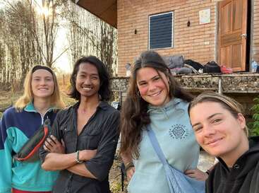 Cuatro amigos están de pie frente a una casa rústica de ladrillo, sonriendo cariñosamente a la cámara. La luz del sol se filtra entre los árboles detrás de ellos, creando un ambiente relajado y alegre.