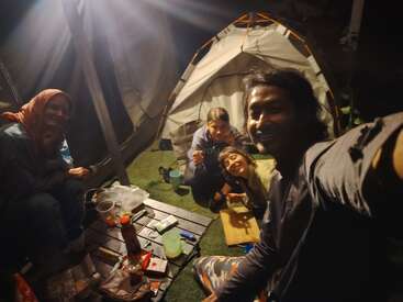 Cuatro amigos están de acampada por la noche, sentados alrededor de una mesa con bocadillos, sonriendo bajo una luz, con tiendas de campaña al fondo, compartiendo juntos un momento alegre.