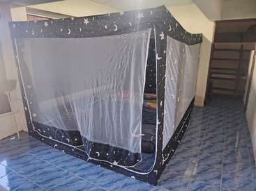 Una gran tienda con mosquitera y dibujos de estrellas y lunas cubre una cama en una habitación alicatada. Cerca hay estanterías, una lámpara y un suelo azul estampado.