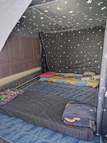 La imagen muestra una acogedora habitación con dos colchones de suelo, ropa de cama de colores y cortinas y techo con motivos de estrellas y lunas, que crean un ambiente de tienda de campaña de ensueño.