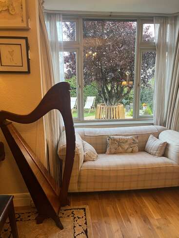 A cozy living room features a plaid sofa with cushions, a wooden harp, framed art, and a large window overlooking a garden with deck chairs.