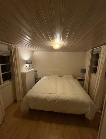 A cozy bedroom with wood-paneled ceiling, light wood flooring, a large bed with white bedding, two windows, bedside tables, lamps, and gentle warm lighting.