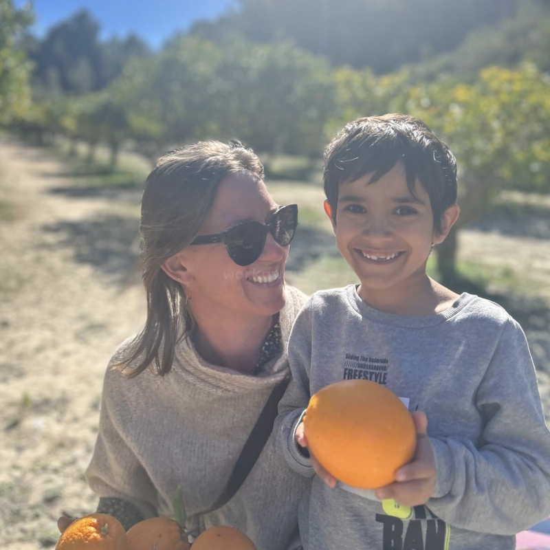 The image depicts a woman and a boy standing together in an orchard, with the boy holding an orange and the woman wearing sunglasses.