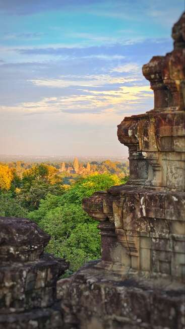 Ancient stone ruins overlook a dense green forest, with distant temple spires rising above the trees under a pastel sky at sunset, evoking tranquility and history.