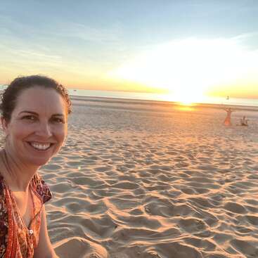 A imagem mostra uma mulher tirando uma selfie em uma praia de areia ao pôr do sol, com o sol se pondo sobre a água ao fundo.