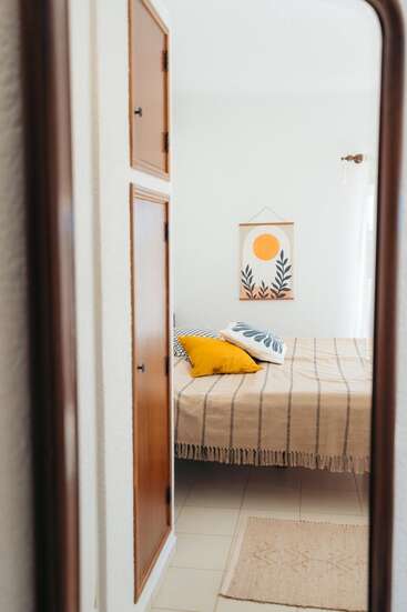 Essa imagem mostra um quarto aconchegante refletido em um espelho, com uma cama com almofadas estampadas, um cobertor listrado, um tapete, armários de madeira e arte minimalista nas paredes.