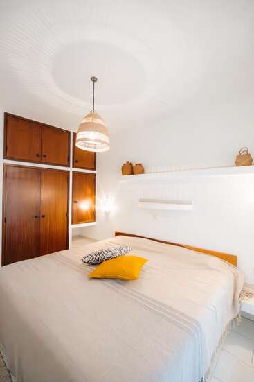 Esse quarto aconchegante tem uma cama grande com roupa de cama bege, almofadas decorativas, armários de madeira, um pendente de luz trançado e prateleiras minimalistas com cerâmicas decorativas.