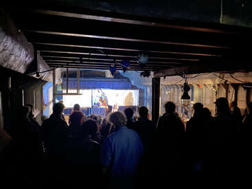 A crowd gathers in a dimly lit, industrial-style venue, facing a small stage where performers, illuminated by bright lights, entertain the attentive audience.