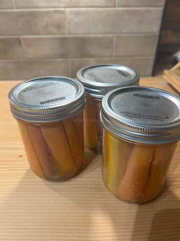 Three glass jars with metal lids, filled with whole carrots preserved in liquid, sit on a wooden surface in a kitchen setting with tiled background.