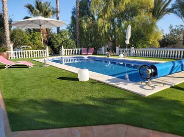 Un hermoso patio trasero cuenta con una piscina azul resplandeciente, exuberante césped artificial, tumbonas, palmeras, sombrillas y barandilla blanca de estilo mediterráneo, que evocan la relajación y el lujo.