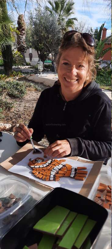 A smiling woman sits outdoors, wearing a black hoodie and sunglasses, happily creating a tiger mosaic artwork with colorful tiles, surrounded by lush greenery and sunlight.