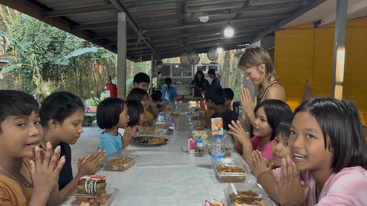 Eine Gruppe von Kindern und ein Erwachsener sitzen an einem langen Tisch unter einem überdachten Außenbereich, lächeln und drücken ihre Handflächen vor dem Essen aneinander.