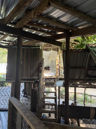 Dieses Bild zeigt ein rustikales Holzhaus mit einem Metalldach, offenen Wänden, einer Holztreppe, einem gemütlichen oberen Bett mit Moskitonetz und darunter sitzenden Menschen.