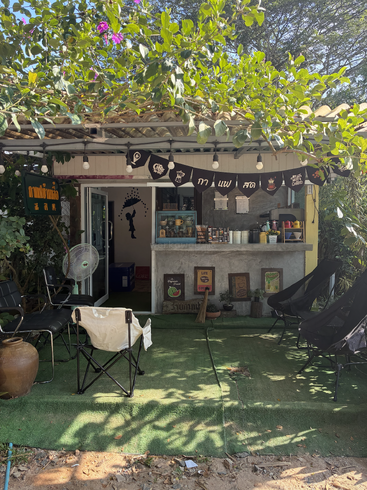 Ein kleines, gemütliches Außencafé mit grünem Kunstrasen, schwarzen Stühlen, hängenden Lichtern, belaubten Bäumen, bunten Schildern und einem Wandgemälde eines Mädchens mit einem Regenschirm.