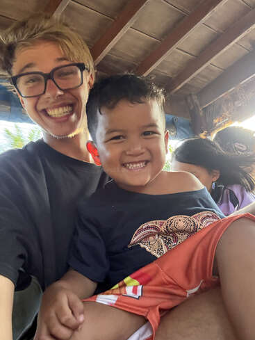 A young man wearing glasses and a child are smiling widely while sitting together. They seem happy and joyful under a wooden roof, enjoying the moment.