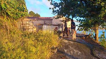 Two people stand on a sunlit, rustic terrace near a small house, surrounded by greenery and trees, overlooking blue water, evoking peaceful, nature-filled vibes.