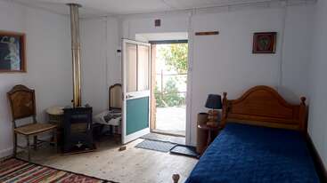 This cozy room features a wood stove, two chairs, a single bed with blue bedding, a colorful rug, a lamp, and an open door to nature.
