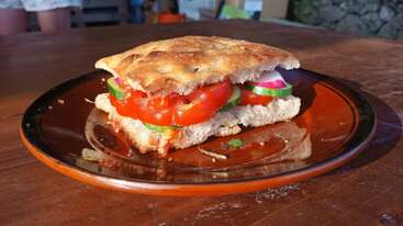 Un sandwich frais est posé sur une assiette brune, rempli de tranches de tomate, de concombre et d'oignon rouge, le tout entre deux morceaux de pain focaccia rustique et doré.