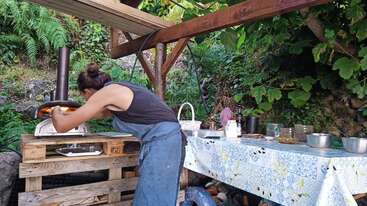 Une personne en tablier prépare une pizza dans un four à bois en plein air, entouré de verdure, avec divers ingrédients et ustensiles sur une table couverte à proximité.