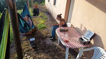 Deux personnes se détendent en plein air ; l'une est assise à une table recouverte d'une nappe à carreaux rouges, tandis que l'autre se prélasse dans un hamac. La lumière du soleil projette de longues ombres dans l'après-midi.