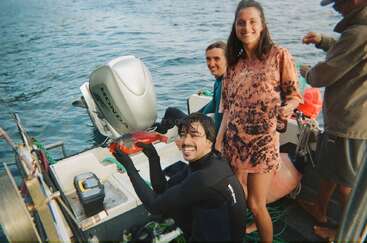 Quatre personnes se trouvent sur un bateau, souriant et s'amusant. L'une d'entre elles, vêtue d'une combinaison de plongée, brandit fièrement un poisson rouge au bord de l'eau, célébrant ainsi sa prise.