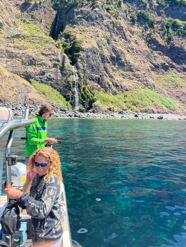 Deux personnes pêchent depuis un petit bateau dans une eau bleue et limpide. Derrière eux, une falaise rocheuse abrupte présente une végétation verdoyante et une cascade. Un paysage magnifique.