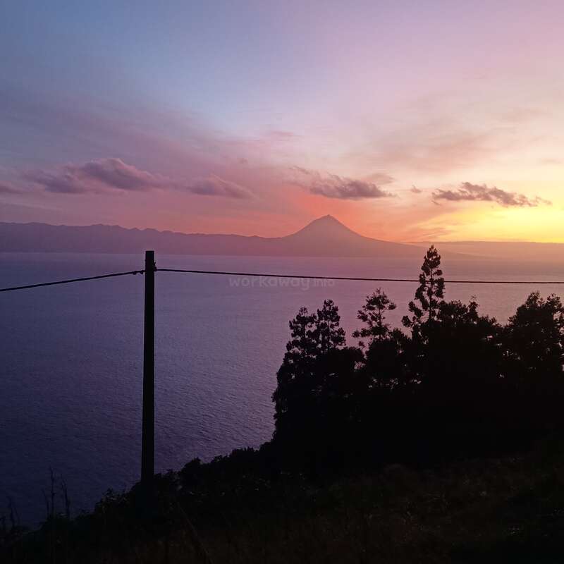 A stunning sunset with vibrant purple and orange hues, overlooking calm ocean waters, silhouetted trees, a volcano in the distance, and a simple power line crossing.