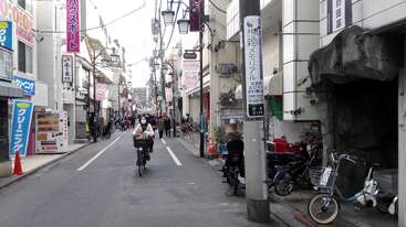 A bustling Japanese street scene with people walking and cycling, shops on both sides, signs in Japanese, vending machines, and bicycles parked along the sidewalk.