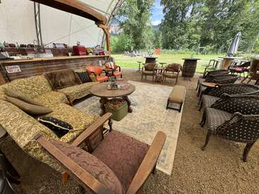 This cozy outdoor seating area features mismatched vintage furniture, a large rug, and a coffee bar, surrounded by greenery and trees, creating a relaxed atmosphere.