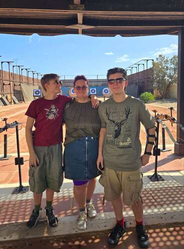 Tres personas permanecen juntas, sonriendo bajo una estructura sombreada en un campo de tiro con arco al aire libre. Las dianas se ven al fondo, y todos llevan ropa informal de verano y gafas de sol.