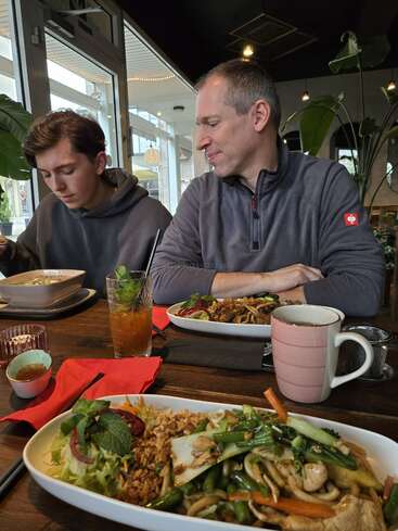 Dos hombres, probablemente padre e hijo, se sientan juntos a la mesa de un restaurante. Están disfrutando de platos de cocina asiática, tragos y bebidas calientes, compartiendo una comida.