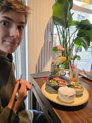 Una persona joven se sienta en una mesa de madera junto a la ventana, con los palillos en la mano. En una bandeja decorativa se sirve deliciosa comida asiática, fideos y bebidas frescas. Cerca, exuberantes plantas.