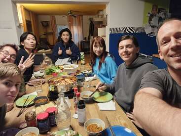 Un grupo de siete amigos están reunidos alrededor de una mesa disfrutando de una comida juntos, sonriendo, haciendo signos de paz y posando felices para un selfie. Ambiente acogedor.