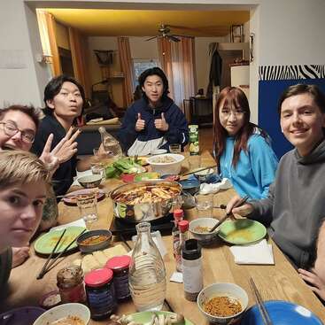 Un grupo de siete amigos están reunidos alrededor de una mesa disfrutando de una comida juntos, sonriendo, haciendo signos de paz y posando felices para un selfie. Ambiente acogedor.