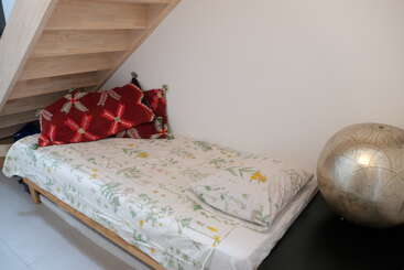 Esta imagem mostra uma cama pequena com roupa de cama floral sob uma escada de madeira. Almofadas com estampas vermelhas e uma luminária metálica decorativa ficam próximas a uma parede simples.