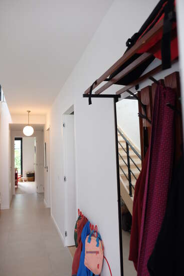 Um corredor moderno e bem iluminado apresenta paredes brancas, um cabide, mochilas coloridas, uma escada com um corrimão preto, portas fechadas e uma luz de teto redonda.