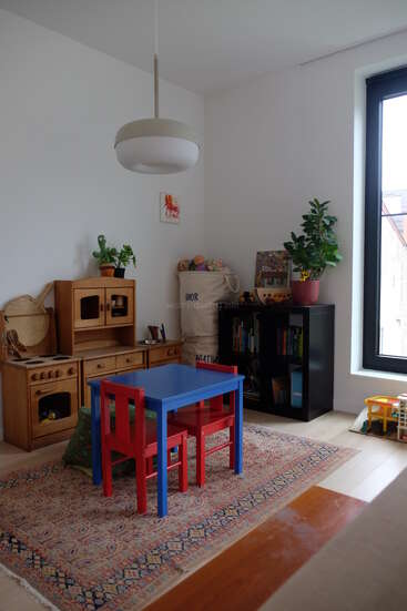 Uma aconchegante sala de jogos para crianças apresenta uma mesa azul com cadeiras vermelhas, uma cozinha de madeira para brincar, estante, plantas, tapete colorido, janela grande e vários brinquedos.