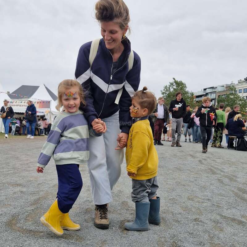 Una mujer sonríe mientras da la mano a dos niños con la cara pintada. Llevan botas de colores, disfrutando de un evento al aire libre con mucha gente de fondo.