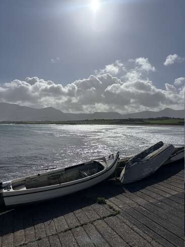 Two small, weathered boats rest on a concrete shore. The sun shines brightly above, illuminating shimmering water, distant green fields, mountains, and fluffy clouds.