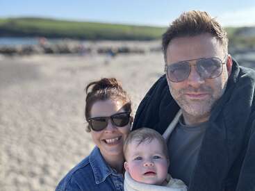 A happy family of three, wearing sunglasses, poses for a selfie at a sunny beach with sand, blue sky, and green hills in the background.