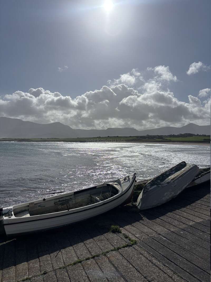 Two small, weathered boats rest on a concrete shore. The sun shines brightly above, illuminating shimmering water, distant green fields, mountains, and fluffy clouds.