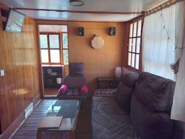 Das Bild zeigt ein gemütliches Wohnzimmer mit Holzvertäfelung, Couch, Couchtisch und Sessel, mit einem Fenster und Fernseher an der linken Wand und einer Tür zu einem anderen Raum an der Rückwand.